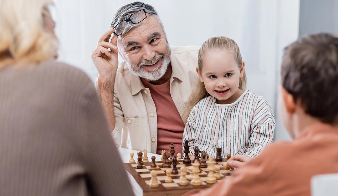Ein Seniorenpärchen sitzt mit seinen Enkelkindern glücklich im Wohnzimmer und spielt Schach als Symbolbild für strategisch kluge Entscheidungen bei Investments für die Altersvorsorge | Wohnen im Alter